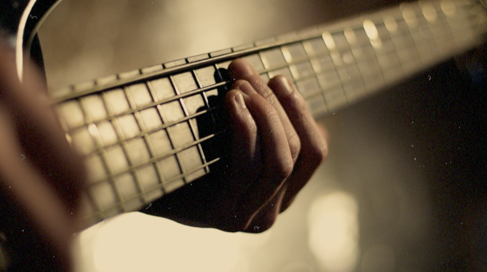 A close-up image of fingers playing on the neck of a 5-string bass guitar. The background is out of focus, and the image has a grainy tan tint to it.