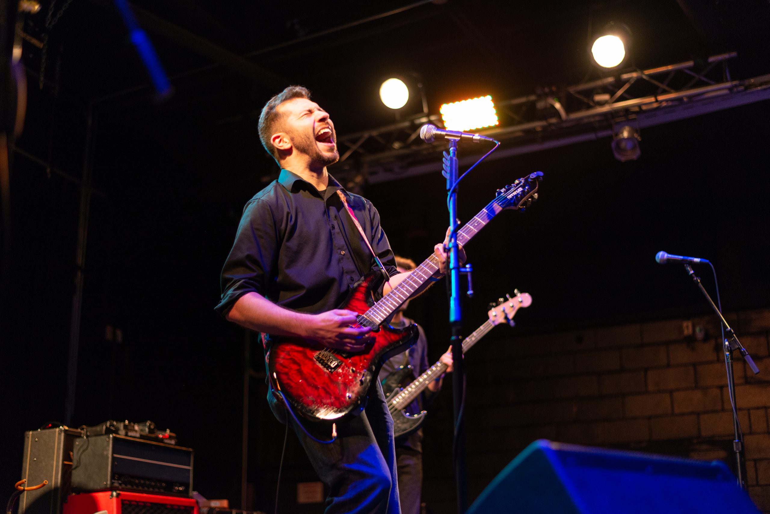 Bryce Kalal singing and playing guitar on stage at The Garage. He has pulled away from the mic to sing a high note with his eyes shut and his mouth wide open.