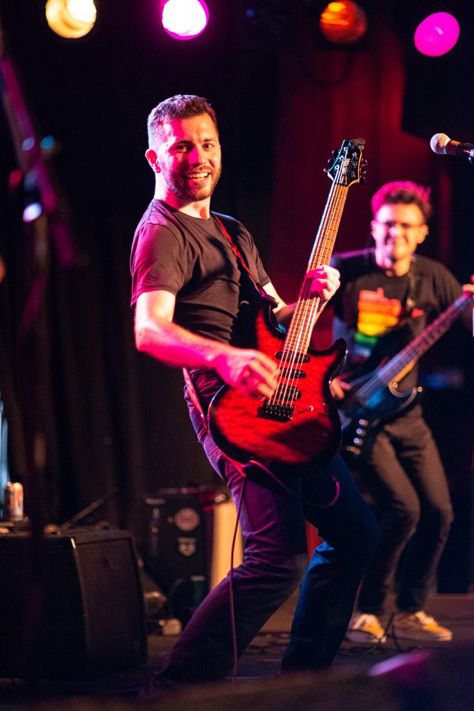 Bryce holding out a note on the guitar and smiling. Alec is playing the bass in the background, also smiling.